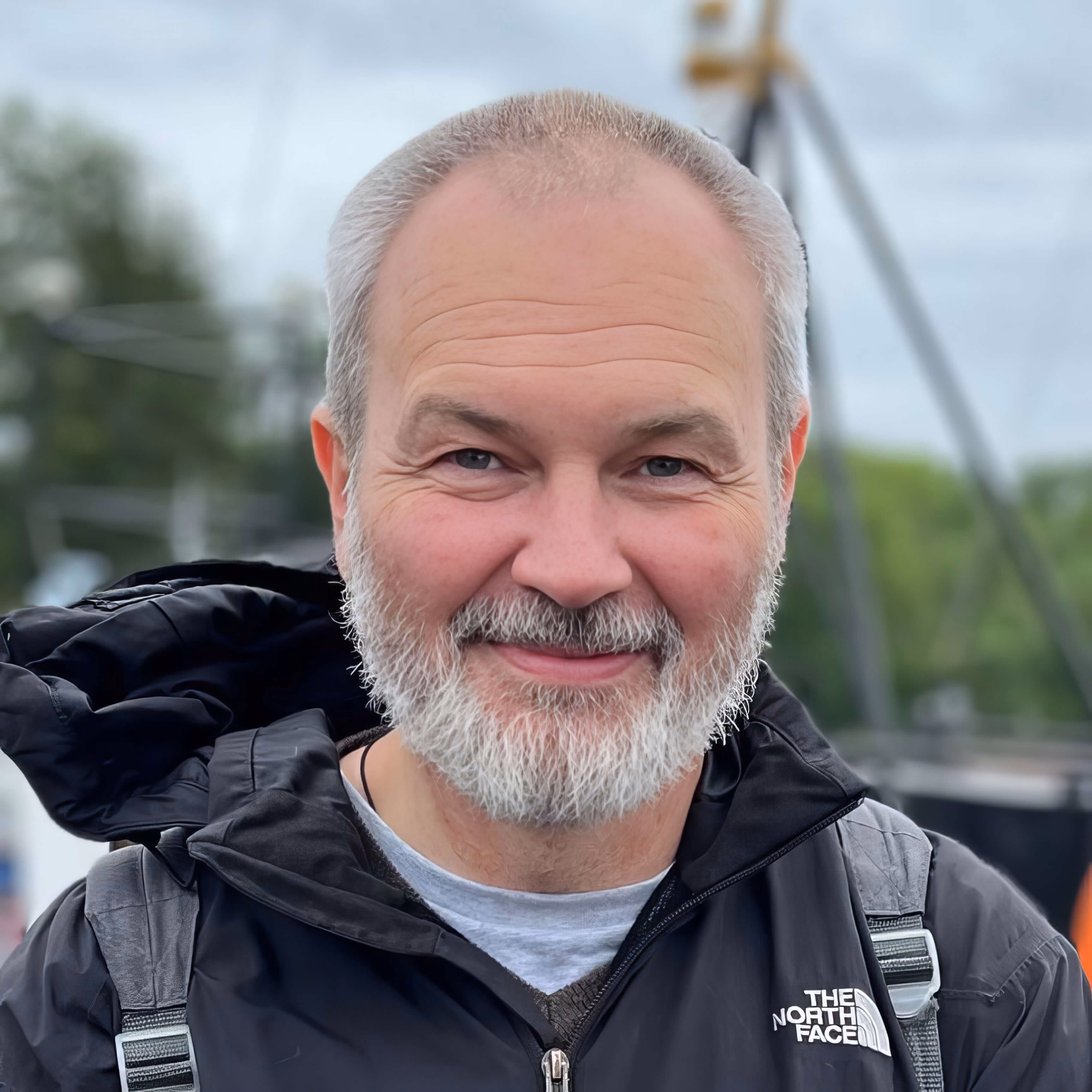 Детальный портрет Виктора Викторовича Аверкиева (дата рождения 1973-12-09) крупным планом. Мужчина европеоидной внешности, на вид 50-52 года. У него очень короткая стрижка (ежик), волосы серебристо-седые. Борода густая, аккуратно подстриженная, седая с вкраплениями темных волос в области усов. Лоб высокий, с заметными глубокими горизонтальными морщинами. Глаза светло-серо-голубые, в уголках глаз сетка морщин. На щеках и носу здоровый румянец. Взгляд прямой, выражение лица спокойное, располагающее, с легкой полуулыбкой. Одет в черную куртку бренда The North Face, на плечах лямки рюкзака. Задний план размыт, угадываются очертания мачт или конструкций на открытом воздухе.